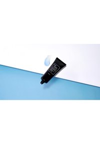 Tube noir de crème, avec une finition mate et un capuchon à charnière, reposant sur une surface bleue. Une petite goutte de crème bleu clair est à proximité.