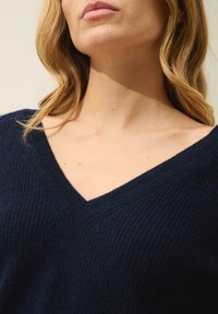 Granatowy sweter z dekoltem w kształcie litery V, teksturowana dzianina oraz subtelne poziome linie. Zbliżenie skupione na dekolcie i ramionach.