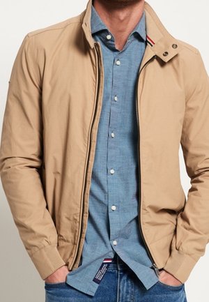 Mężczyzna w beżowej bluzie na zamek, pod którą ma niebieską koszulę na guziki, oraz w niebieskich dżinsach, z rękami częściowo w kieszeniach bluzy.