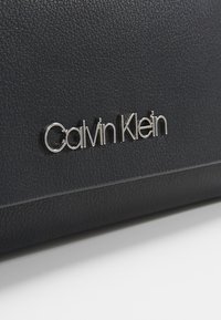 Černý texturovaný kožený povrch se stříbrným kovovým logem značky "Calvin Klein" připevněným.