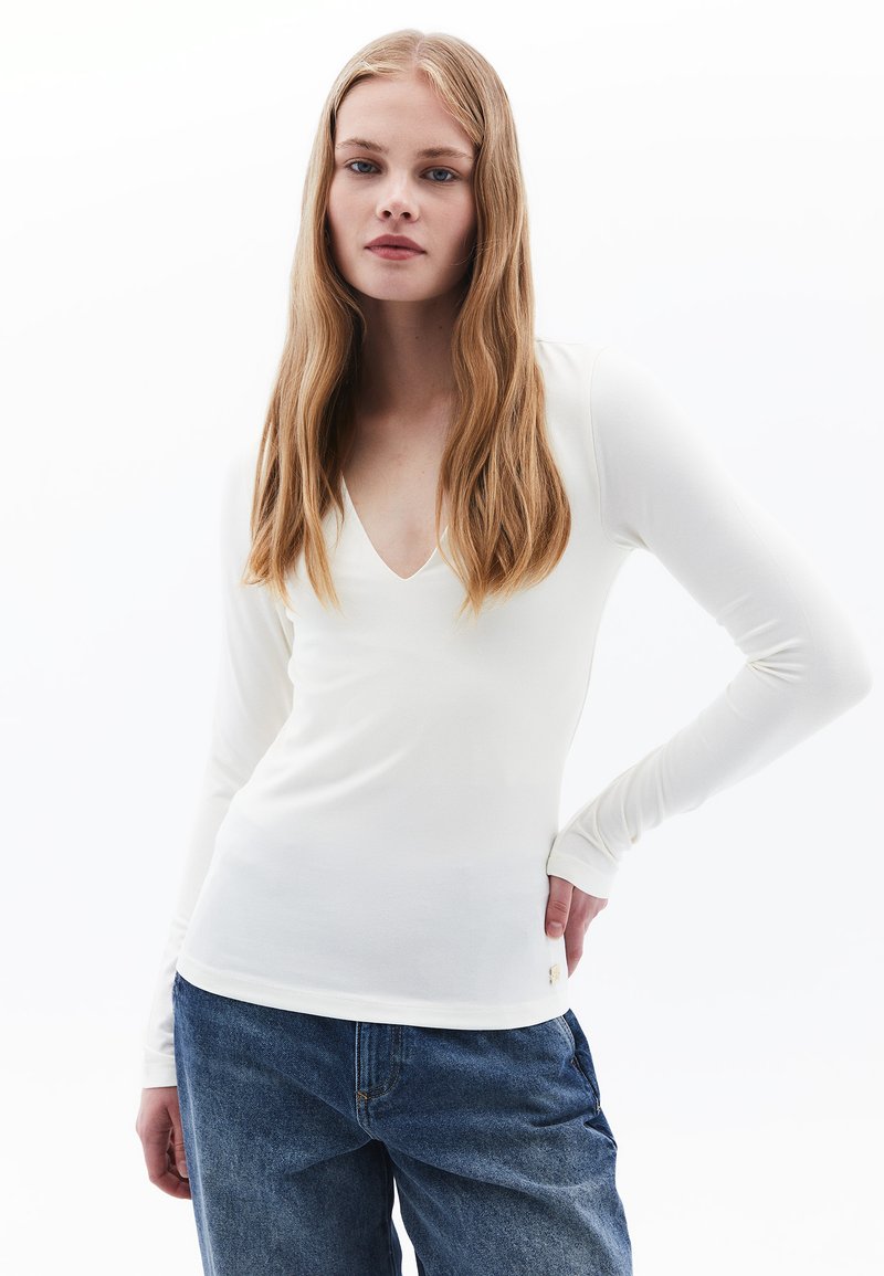 Maglietta a maniche lunghe e scollatura a V realizzata in morbido tessuto bianco con una silhouette aderente. Abbinata a jeans in denim blu morbidi.
