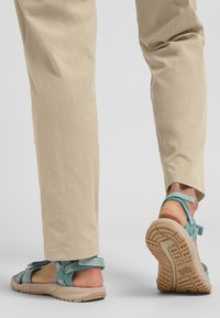 Turquoise sandals with textured straps and adjustable closures, worn with light beige pants. Durable rubber sole with traction pattern.