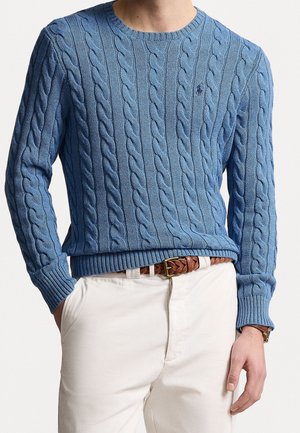 Mand iført blå kabelstrikket sweater, hvide bukser med brunt flettet læderbælte, stående med den ene hånd i lommen mod en lys baggrund.