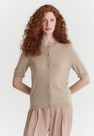 Mujer con cabello largo rizado y rojo lleva un suéter beige de manga corta con botones y pantalones sueltos de color rosa claro, de pie con las manos detrás de la espalda.