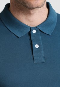 Türkisen Poloshirt mit Kragen, drei weißen Knöpfen und einem strukturierten Stoff. Nahaufnahme hebt die Nähte und das Kragen-Design hervor.