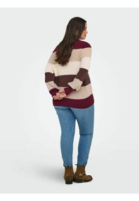Frau mit langen dunklen Haaren, die einen gestreiften Pullover, blaue Jeans und braune Knöchelstiefel trägt, steht mit hinter dem Rücken verschränkten Händen.