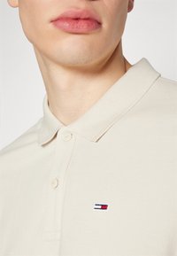 Beige Poloshirt mit geripptem Kragen, zwei Knöpfen und einem kleinen gestickten Logo mit roten, weißen und blauen Streifen auf der Brust.