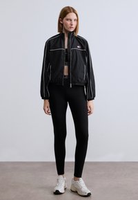 Μαύρο μπουφάν ανεμοφράκτη με λευκές ραφές, φορεμένο πάνω από μαύρο crop top και κολάν. Το μοντέλο στέκεται με αθλητικά παπούτσια σε ουδέτερο πάτωμα.