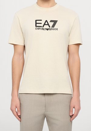 Mężczyzna ubrany w beżowy T-shirt z krótkim rękawem z czarnym logo „EA7 Emporio Armani” oraz jasnobrązowe spodnie, stojący na jednolitym tle.