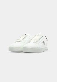 Baskets blanches en cuir avec un bout rond, semelle plate blanche, accents gris clair et détail du logo noir sur le talon et la languette.
