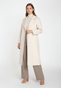 Donna in piedi che indossa un lungo cappotto crema su un dolcevita beige, pantaloni larghi taupe e stivaletti con tacco color tan contro uno sfondo bianco.