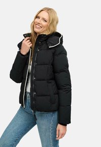 Chaqueta de plumas negra con capucha, con un ribete blanco, textura acolchada, botones a presión y bolsillos laterales. Usada sobre una camiseta blanca y jeans.