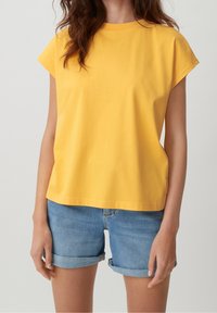 Kobieta ubrana w gładką żółtą bluzkę z krótkim rękawem i podwinięte niebieskie dżinsowe szorty, stojąca na tle jasnego tła.