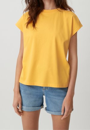 Kvinna i en enkel gul kortärmad topp och rullade blå denimshorts, stående mot en enkel ljus bakgrund.