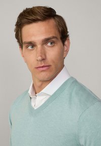 Maglione azzurro chiaro a collo a V con una trama liscia, indossato sopra una camicia bianca con colletto. Il modello ha i capelli corti di colore marrone e guarda leggermente di lato.