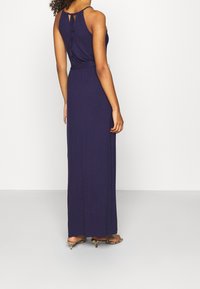 Mörkblå maxiklänning med ärmlös design, knytning på baksidan och en flödande silhuett. Materialet verkar mjukt och lätt.