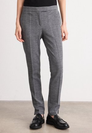 Personne portant un pantalon slim gris à motif chevrons, associé à des mocassins en cuir noir, debout sur un sol neutre.