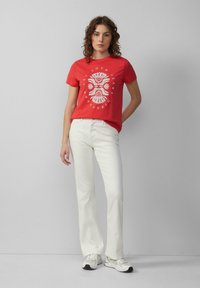 T-shirt en coton rouge avec un imprimé graphique blanc sur le devant, associée à un jean blanc et des baskets légères. Le modèle se tient contre un fond uni.