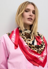 Pañuelo de seda con estampado de leopardo y acentos en rojo y nude, un logo audaz y un detalle de cadena, colocado con estilo alrededor del cuello.