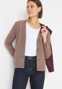 Braune, strukturierte Strickjacke mit geripptem Kragen und Knopfverschluss, kombiniert mit einem weißen T-Shirt und dunklen Jeans. Auffällige Details sind sichtbare Nähte.