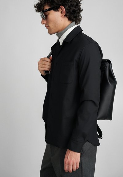 Camisa negra de botones con un bolsillo delantero, combinada con un suéter de cuello alto gris y pantalones grises. El modelo sostiene una mochila negra.