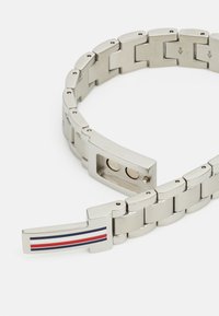 Pulsera de metal plateado con eslabones entrelazados, que presenta un cierre rectangular decorado con franjas rojas, blancas y azules. Textura lisa.