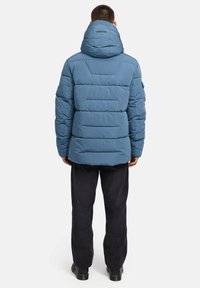 Plavi puffer kaput s visokim kapuljačom, kviltanim dizajnom i elastičnim manžetama. Uparen s tamnim kordon hlačama i crnim čizmama.