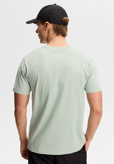 Uomo di spalle che indossa una maglietta a maniche corte verde chiaro e un cappellino da baseball nero su uno sfondo semplice.