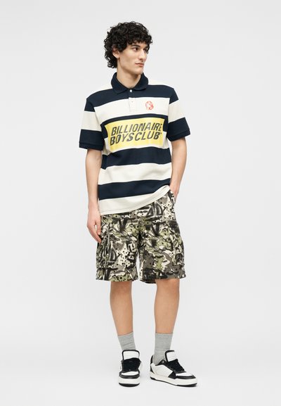 Νέος άνδρας που φοράει μπλε ναυτικό και λευκό ριγέ πόλο με κείμενο "Billionaire Boys Club", καμουφλαρισμένη βερμούδα cargo, γκρι κάλτσες και μαύρα-άσπρα αθλητικά παπούτσια.