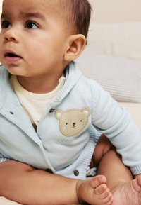 Cardigan azzurro chiaro lavorato a maglia con toppa orso, abbottonato, sopra una camicia color crema. Piedi nudi di un bambino visibili, seduto su una superficie morbida.