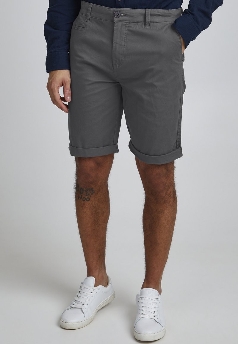 Gray cotton shorts with rolled cuffs, front pockets, and a button closure, paired with white sneakers on tanned legs featuring a tattoo.