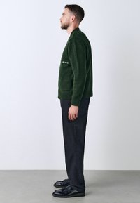 Green knitted cardigan with a textured finish, side pockets, and button closures, worn with dark pants and black shoes.