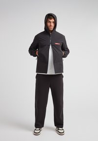 Giacca nera con cappuccio e zip frontale, con un logo rosso. Indossata sopra una maglietta bianca, abbinata a pantaloni neri e sneakers chunky.