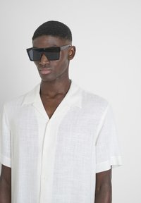 Camisa de lino blanca de manga corta con un sutil patrón texturizado, combinada con grandes gafas de sol rectangulares negras con montura angular y audaz.