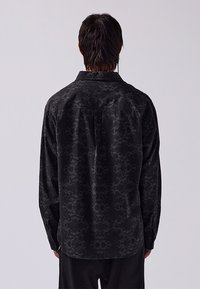 Maglietta nera con una delicata trama a motivi, maniche lunghe e colletto classico. Presenta polsini con bottoni e una cucitura centrale dritta sul retro.
