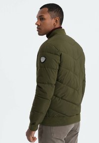Olivgrön pufferjacka med dragkedja i kragen, quiltad textur och ribbade muddar. Har en logotyp-patch på vänster axel.