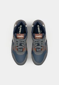 Adidași gri și bleumarin cu o suprafață texturată, având un design perforat, șireturi gri și accente maro pe toc și limbă.