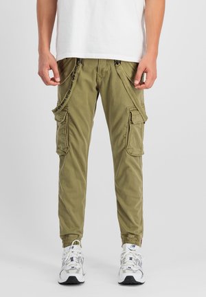 Man die olijfgroene cargobroek met tekstbanden en witte sneakers draagt, staand tegen een effen lichte achtergrond.