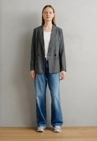Blazer grigio con motivo a zigzag e due bottoni, indossato su una camicia bianca, abbinato a jeans larghi blu chiaro e sneakers grigie.