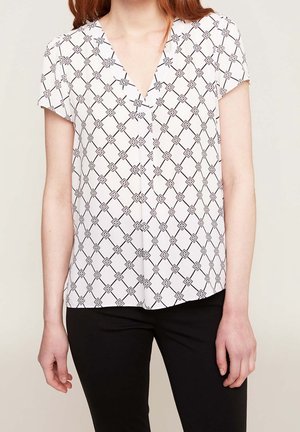 Mujer con blusa blanca de manga corta con patrón geométrico de diamantes negros y pantalones negros, de pie frente a un fondo claro y liso.