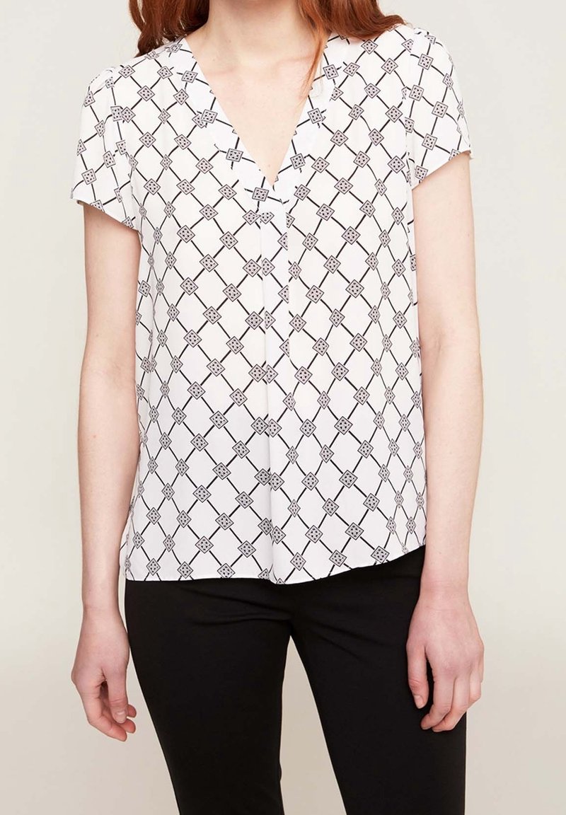 Femme portant un chemisier blanc à manches courtes avec un motif géométrique en losanges noirs et un pantalon noir, debout devant un fond clair uni.