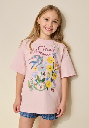Young girl smiling, wearing a pink T-shirt with colorful floral and bird designs and "Fleurs d'Amour" text, paired with blue denim shorts.