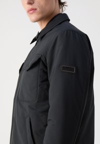 Schwarze isolierte Jacke mit Druckknopfkragen, großer Brusttasche, Seitentaschen und Barbour International-Patch auf dem oberen Arm.