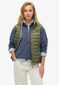 Olivgrön pufferväst med dragkedja, bärs över en blå sweatshirt och en randig skjorta med krage. Krämfärgade byxor fulländar outfiten.