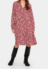 Punainen kukkamekko, jossa on pitkät hihat, v-pääntie ja kerrostettu hame. Valmistettu kevyestä kankaasta. Yhdistetty polven ylle ulottuviin mustiin saappaisiin.