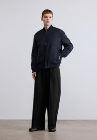 Ναυτικό μπουφάν bomber με φερμουάρ μπροστά, ριμπ γιακά και δύο πλαϊνές τσέπες. Φορεμένο πάνω από άνετο μαύρο παντελόνι και λείες παπούτσια.