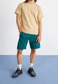 T-shirt di cotone beige con piccolo logo, abbinato a pantaloni da ginnastica teal. Indossato con sneakers nere e calzini bianchi con righe azzurre chiare.