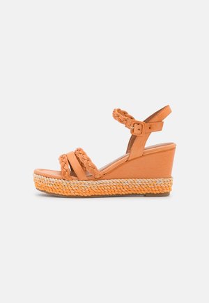 Tan kile sandal med flettede stropper, ankelspænde og vævet platformsål på en lys grå baggrund.