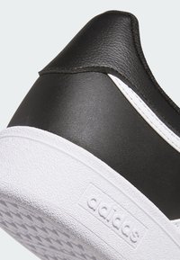 Zapatilla negra con parte superior de cuero texturizado, suela blanca y suela de goma. Presenta acentos cosidos y el logo de "adidas" en el lado.