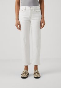 Jeans de mezclilla blanca con corte recto, bolsillos frontales y remaches de cobre, combinados con mocasines texturizados y con patrón en negro y beige.